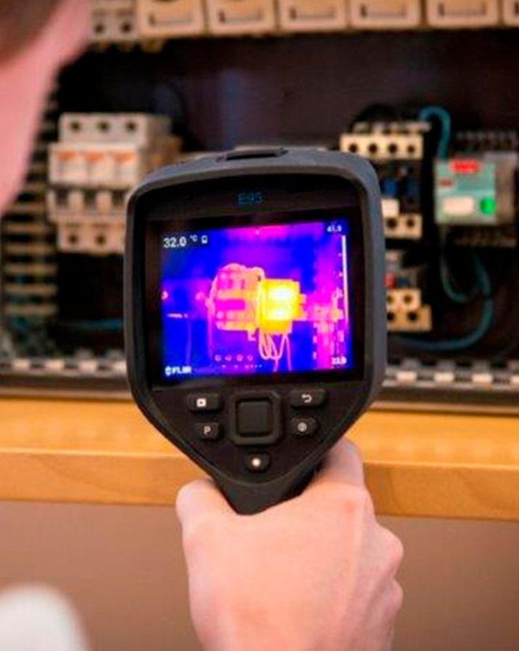 En hånd holder et termografisk FLIR-kamera op mod en eltavle.